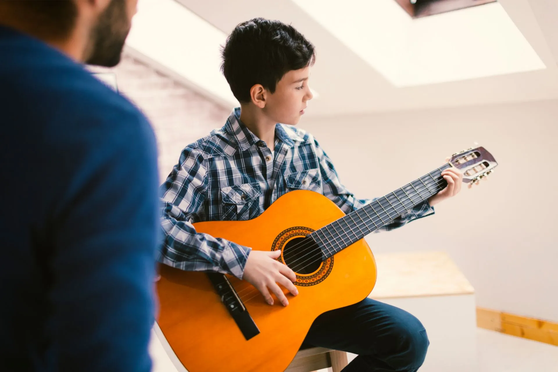 Young boy in plaid shirt learning classical guitar with teacher’s guidance – music lessons in bright, modern room.
