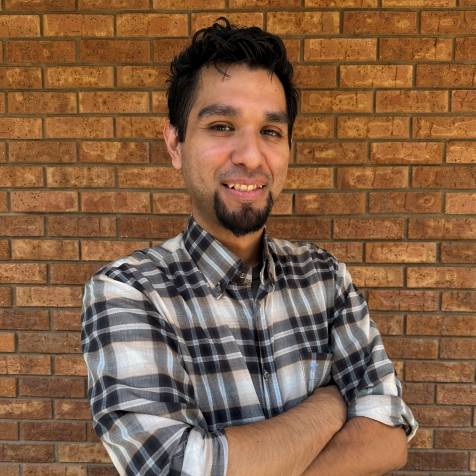 A man wearing a black, white, and gray plaid shirt stands smiling with arms crossed in front of a brick wall. He has short, dark hair and a goatee.