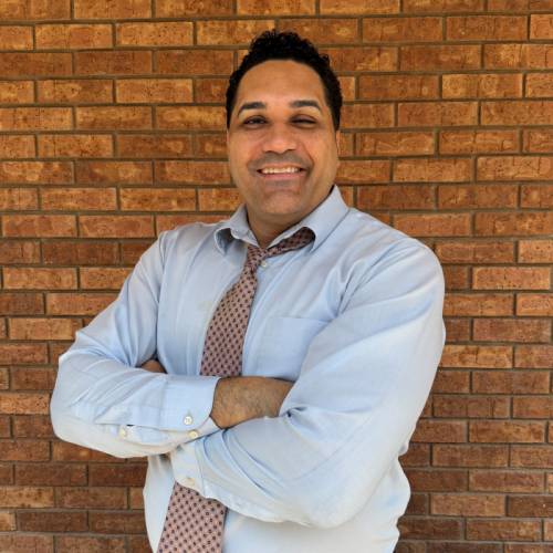 A man wearing a light blue dress shirt and patterned tie stands smiling with arms crossed in front of a brick wall.