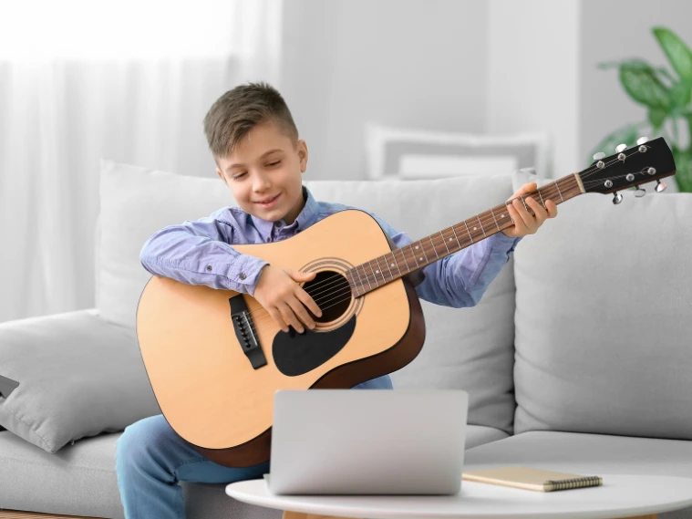Kid interacting with a virtual music class on screen holding a guitar, following along with digital activities and rhythm exercises during an online group lesson