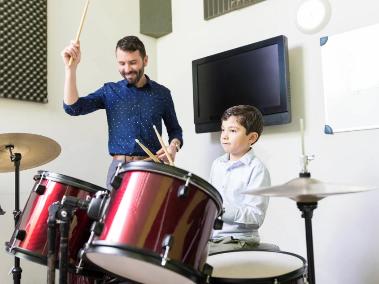 Beginner drummer practicing on a drum set with an instructor guiding proper technique in a music studio