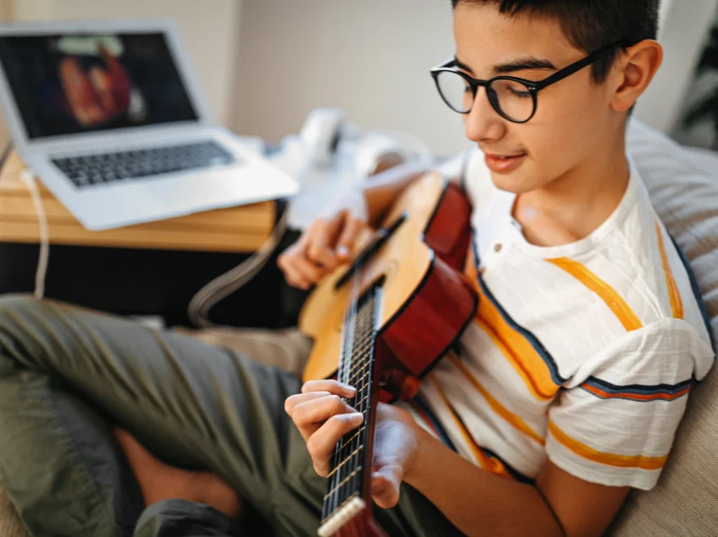 Young student practicing guitar at home while following an online beginner guitar lesson on a laptop